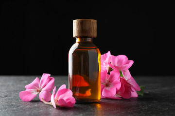 Bottle of geranium essential oil and beautiful flowers on black table, closeup