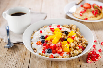 bowl of granola with yogurt and fresh fruits, coffee cup on wood background