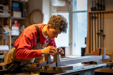 Focused woodworker applies leather pads to bed legs in workshop, enhancing stability and protection. Male craftsman using traditional woodwork methods, handmade furniture, leather work with bed frame