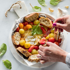 Hands spreading tomato and melted brie cheese onto crusty baguette slices.