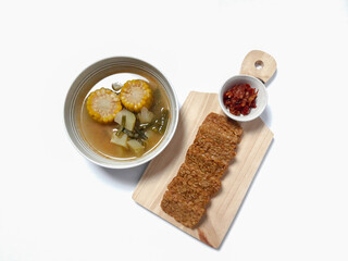 Sayur asam or sayur asem is popular soup in Indonesia. Served in a bowl with fried tempeh and sambal on a cutting board. Isolated white background
