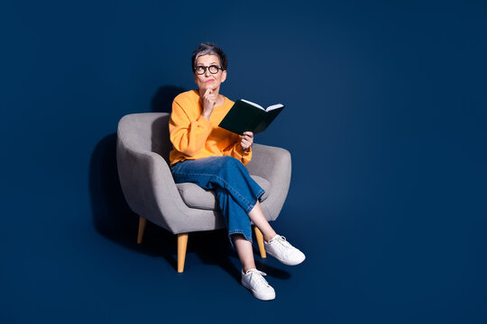 Full length photo of senior female sit soft armchair look empty space read book wear yellow outfit isolated on dark blue color background