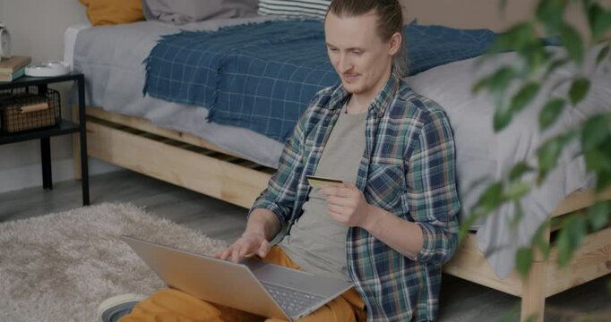 Cheerful young man holding credit card and using laptop computer then expressing joy in bedroom at home. Shopping and banking concept.