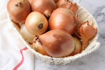 Fresh onions with peels in wicker basket on light marble table, closeup