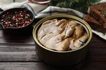 Tasty sprats in tin can, peppercorns, bread and dill on wooden table, closeup