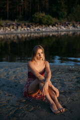 A beautiful young woman in a pink dress is sitting on the seashore at sunset.