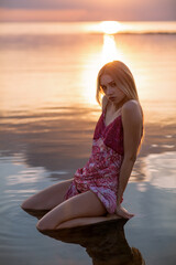 A beautiful young woman in a pink dress is sitting in the water on the seashore at sunset.
