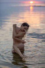 A beautiful woman in lingerie is lying in the water on the seashore at sunset. A girl is hugging herself by the shoulders.