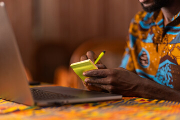 Close-up of an African man's hands writing on a notepad while working on a laptop.
