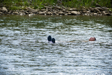 boy swims in a fast mountain river, lies on his back, head and crocs above the water, stones