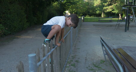Young boy leaning over a wooden fence, not being prudent, wearing a white shirt, with a focused expression, exploring the outdoors, surrounded by greenery on a sunny day