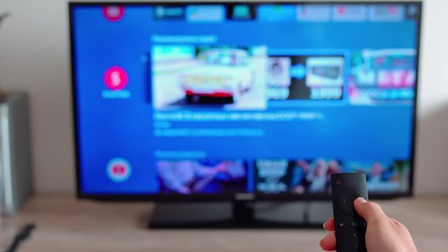 Close-up of female hand holding the TV remote control and flipping through channels on defocused smart tv in living room. Woman indulging in some channel surfing in internet TV. Domestic cinema in 4K