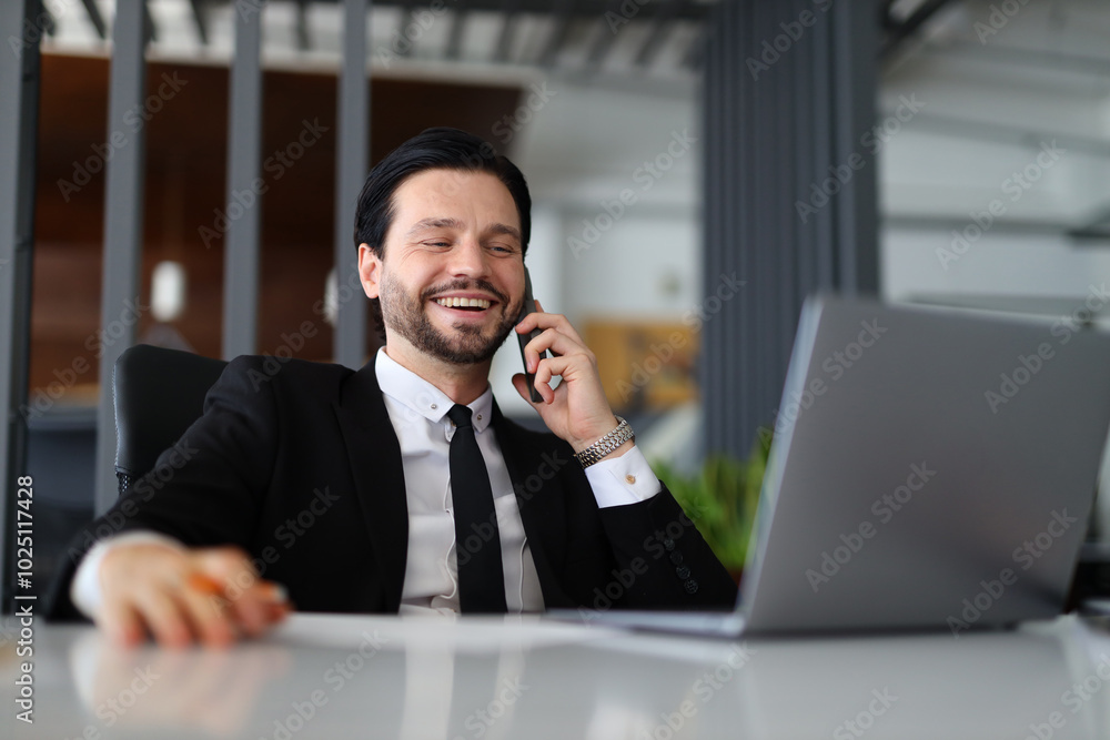 Poster a man is talking on his cell phone while sitting at a desk. he is wearing a suit and tie and he is i
