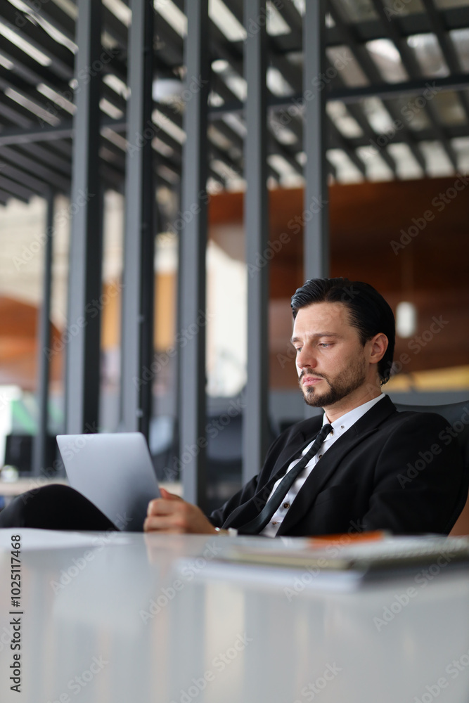 Poster a man in a suit is sitting at a desk with a tablet in front of him. he is reading something on the t