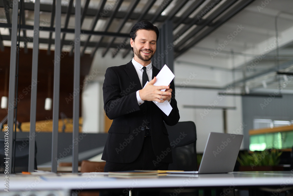 Poster a man in a suit is standing in front of a desk with a laptop and a stack of papers. he is smiling an