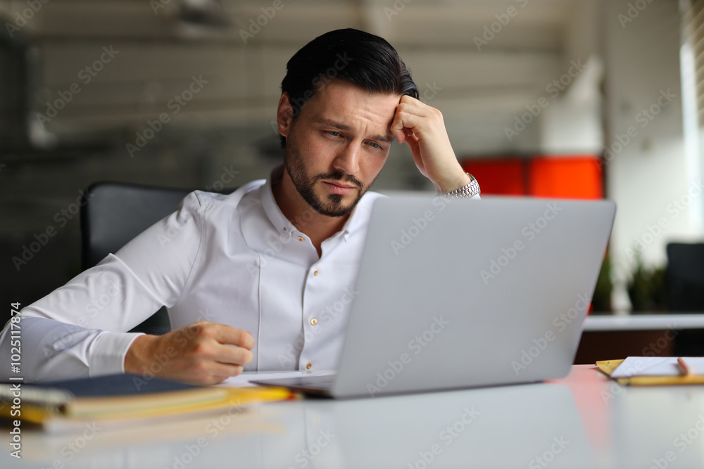 Poster a man is sitting at a desk with a laptop open in front of him. he is looking at the screen with a pu