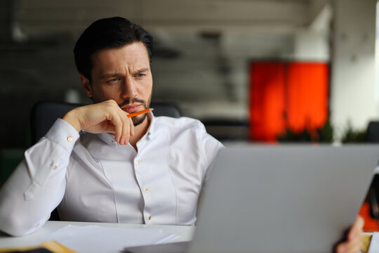 A man is sitting at a desk with a laptop and a pencil. He is looking at the laptop screen and he is deep in thought