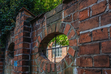 An old brick wall with round openings. In the background a brick building covered in plants is visible.