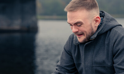 Man With a Short Beard Sitting by the River, Experiencing Deep Emotions on a Chilly Day During the...
