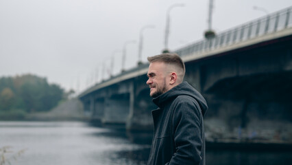 Young Man Enjoying a Quiet Moment by the River Beneath a Bridge on an Overcast Day in Autumn