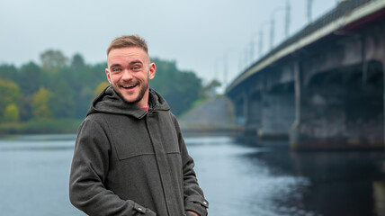 Young Man Smiling by a River Under a Cloudy Sky Near a Bridge in Autumn