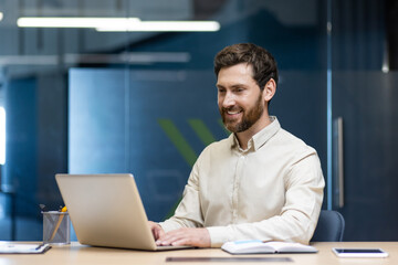 Fototapeta premium A young male businessman works in the office at a laptop, sits at a desk and looks at the screen with a smile