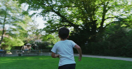 Young boy running through a grassy park, viewed from behind, wearing a white shirt and shorts, surrounded by tall trees and greenery, enjoying an active day outdoors