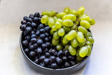 Bunches of black and green grapes lying on a light surface. In the background, a bowl with green and black grapes is visible.