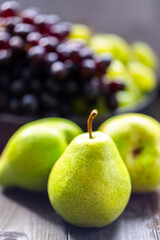 Green pears in the foreground with a blurred background featuring bunches of black and green grapes.