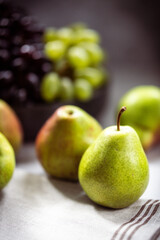 Green pears in the foreground with a blurred background featuring bunches of black and green grapes.
