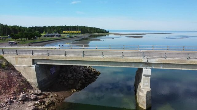 Drone pass between brick and reinforced concrete foundations under a bridge leading to a beach along a calm saltwater bay with a mirror effect reflecting the colors of the sky.