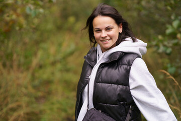 Young beautiful smiling dark-haired girl in vest and hoodie standing against blurred forest background. Concept of unity with nature