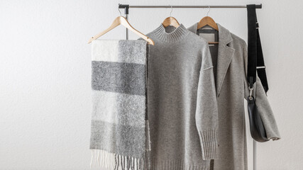 Light grey wool coat or jacket, merino sweater or dress, alpaca scarf and black leather bag on hanger against white wall background. Trendy  monochrome fashion look.