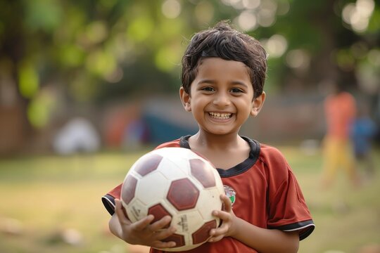Young boy holds soccer ball in park setting. Red shirt, black shorts, vibrant colors. Boy face smiles, happy expression. Soccer ball white with red, black stripes. Park background with trees, fence.
