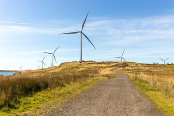 Gravel dirt track road leading to wind farm turbines near freshwater lake in wild grass moor Scottish countryside landscape