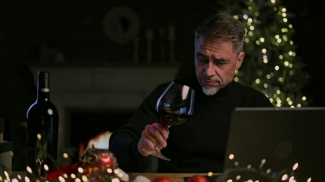 Happy smiling middle aged man at home tasting wine in cosy room with fireplace in winter. Laptop computer, Christmas decor, gift boxes. Concept for web shop ads and vinery holiday offer.