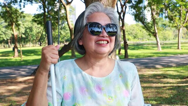 Happiness elderly blind woman optimistic : Portrait elderly blind asian woman sitting on a wooden chair with cane, wearing glasses, sitting alone in a park in the morning, smiling cheerfully.