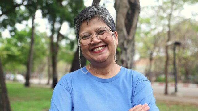 Healthy and cheerful lively retired woman asian standing waving hello looking happy optimist and cheerful looking at the camera for everyone see park with trees and fresh nature good mood.