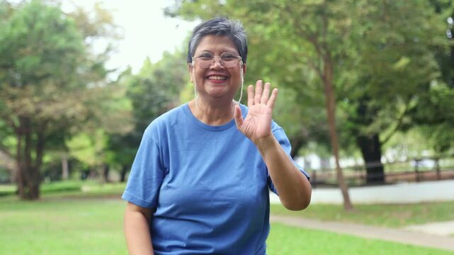 Healthy and cheerful lively retired woman asian standing waving hello looking happy optimist and cheerful looking at the camera for everyone see park with trees and fresh nature good mood.