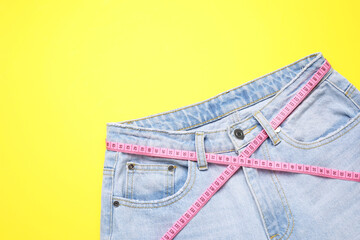 Jeans and measuring tape on yellow background, top view