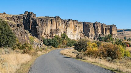 A winding paved road leads towards a dramatic rock formation, with fall foliage in the foreground and a clear blue sky.