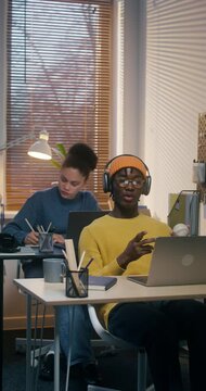 A man and a woman use laptops working at desks in the office. A man in a hat and headphones is chatting on video chat and playing with a baseball
