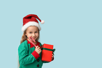Obraz premium Cute little girl in elf costume with gift box and candy cane on blue background
