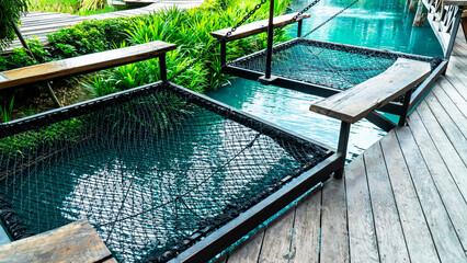 Robe net bed hanging over water river at resort background. Braid string seat or hammock with wood bench beside pond