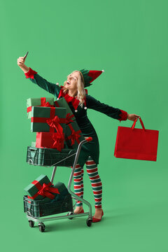 Happy young woman dressed as elf with shopping cart and Christmas gifts taking selfie on green background