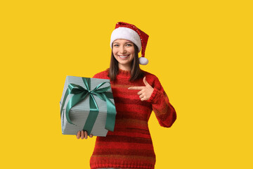 Beautiful young woman in Santa hat pointing at gift box on yellow background