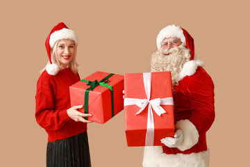 Santa Claus and beautiful mature woman in Christmas hats with gift boxes on brown background