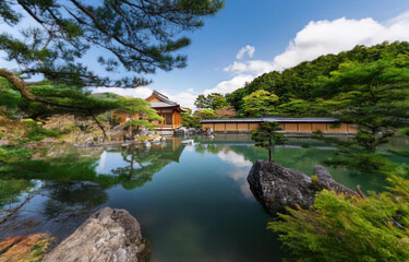 Obraz premium A traditional Japanese wooden structure sits on a small island in the center of a pond surrounded by lush greenery and a blue sky with white clouds.