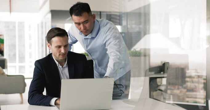 Teammates working on joint on-line project, share opinion and business solutions using laptop. Chinese office employee help with software to mature Hispanic colleague, explain program, help with task