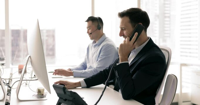 Workflow of helpline center agency, mature office employee sitting at workplace desk with PC holds telephone handset, answers incoming calls from company clients, provide professional support remotely